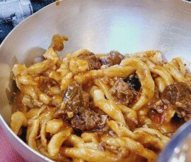 A close-up of thick, hand-rolled Tuscan pasta coated in a rich, chunky meat ragu, served in a deep metal bowl.