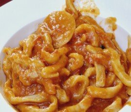 A white bowl filled with thick, hand-rolled pici pasta generously coated in a smooth, orange-red tomato-based sauce, set on a table with a red tablecloth.