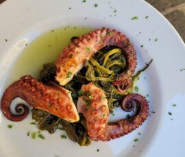 Grilled octopus tentacles served over a bed of cooked green vegetables on a white plate, drizzled with olive oil and chopped parsley.