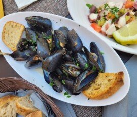 An oval white plate filled with steamed mussels in a light broth, garnished with fresh parsley and served with toasted bread slices.