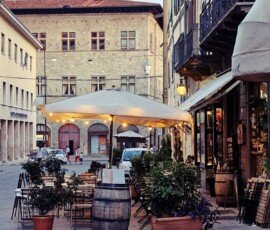 Ogródek restauracyjny na brukowanej ulicy w Pistoi we Włoszech. Drewniane stoliki i krzesła są ustawione pod dużym parasolem z rozwieszonymi lampkami, obok ozdobnej drewnianej beczki po winie. Ulicę otaczają zabytkowe kamienice z okiennicami, a w tle widać spacerujących przechodniów.