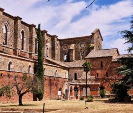 A courtyard view of the medieval Abbey of San Galgano ruins in Tuscany, featuring red brick lower walls, towering stone walls with arched gothic windows, a tall cypress tree, and a small palm tree under a bright blue sky.