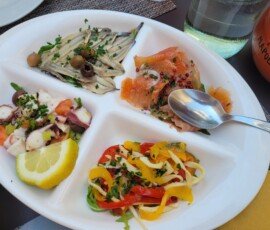 A white divided plate featuring a variety of Italian seafood appetizers: marinated anchovies with olives, salmon carpaccio with pink peppercorns, octopus salad with tomatoes and lemon, and a mixed bell pepper salad.