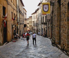 A narrow, sloping street paved with large, uneven stone slabs in Volterra, Tuscany, flanked by tall, old stone and stucco buildings. Pedestrians walk along the street, which features historic flags, a vintage metal streetlamp, and a yellow banner with a red and white crest hanging from a wall.