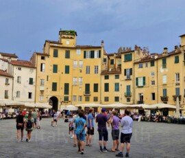 A view of the wide, oval-shaped Piazza dell'Anfiteatro in Lucca, Italy, surrounded by tall, yellow historic buildings with green window shutters. In the center of the stone-paved square, groups of tourists gather, while outdoor restaurant tables with large white umbrellas line the base of the buildings under a dramatic, overcast blue-grey sky.