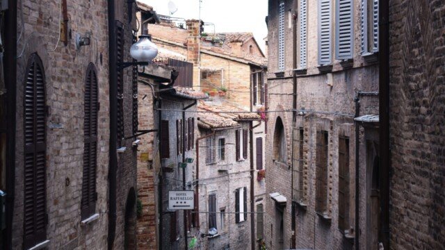 Narrow sloping street between tall brick buildings in Urbino, with a dark car parked on the pavement.