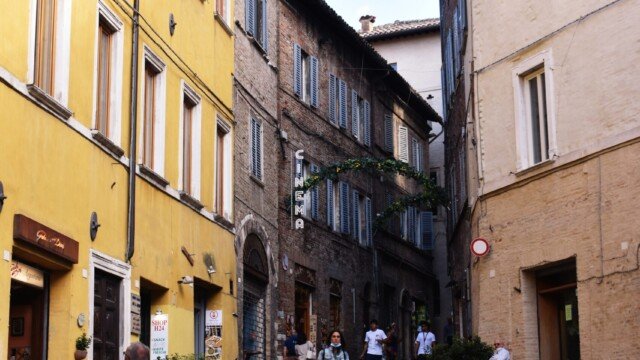 Pedestrians walking along a steep paved street lined with yellow and stone buildings and a cinema sign in Urbino.