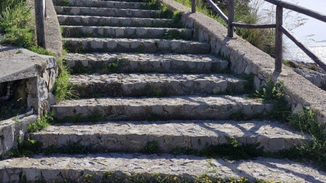 Stone stairs with a wooden railing leading uphill along the Sentiero Apprezzami l'Asino trail in Sapri, Campania, Italy.
