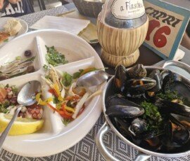 A multi-compartment white ceramic dish containing marinated anchovies, an octopus and tomato salad with lemon, and a squid salad with peppers, served next to a small metal pot filled with steamed mussels at La Sbirra restaurant. La Sbirra Termoli review.