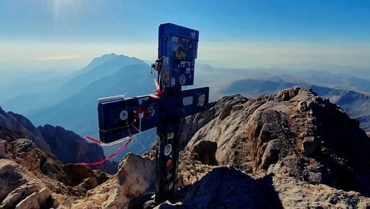 Summit cross covered in stickers and ribbons at the top of Corno Grande overlooking the rugged mountain ridge and surrounding valleys in Gran Sasso, Abruzzo, Italy.