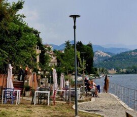 Lakeside promenade in Piediluco with outdoor cafe tables, people relaxing, and a view across the water toward Labro on the distant hill.