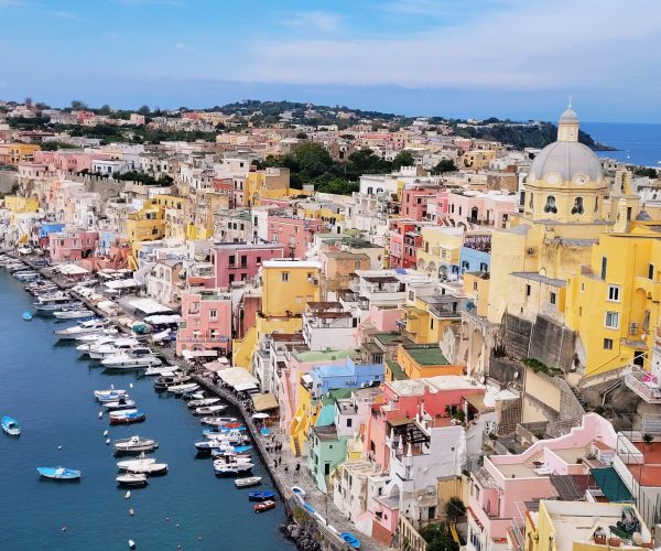 yellow, blue, white and orange houses stand in a port by the sea, where you can see boats. Behind the houses you can see the sea in Procida Island Italy