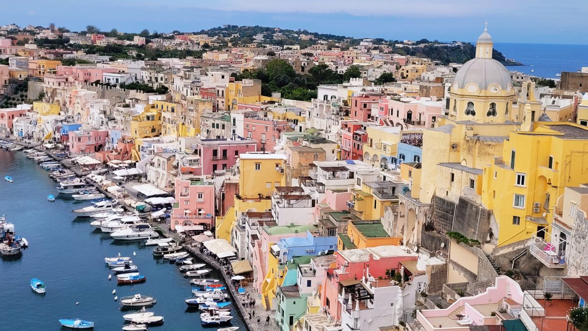 yellow, blue, white and orange houses stand in a port by the sea, where you can see boats. Behind the houses you can see the sea in Procida Island Italy