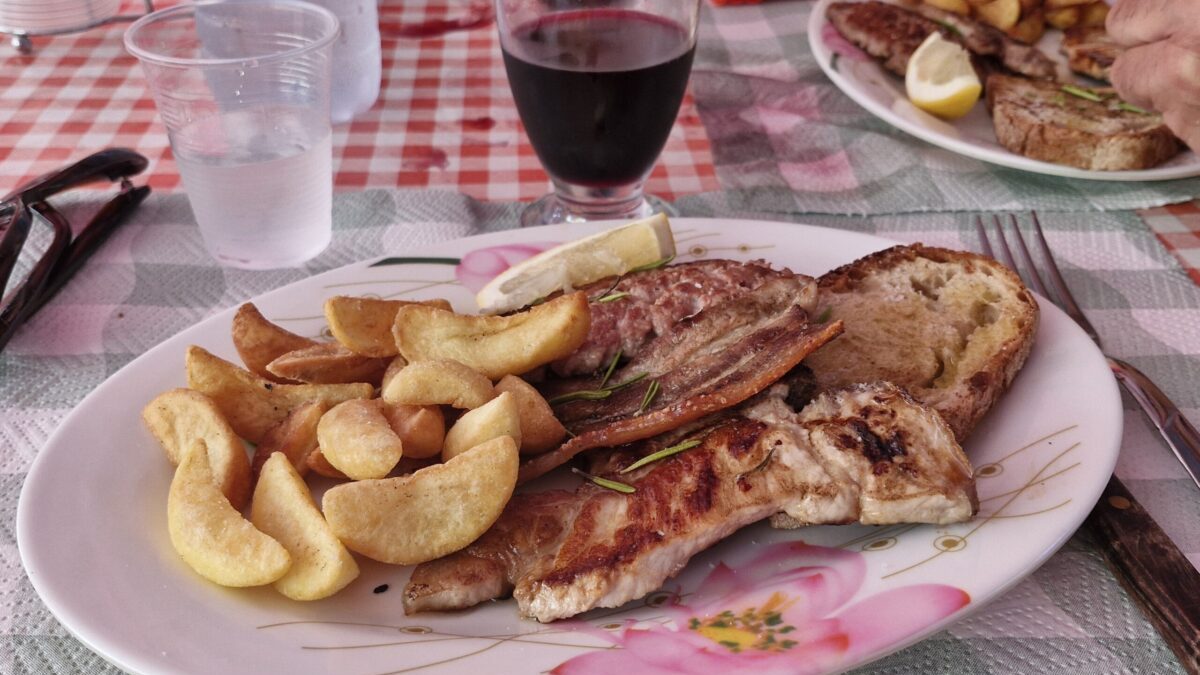 Grillowane mięso z frytkami i pieczywem, podane na plastikowym talerzu w restauracji Punto di Ristoro La Cascata przy Cascata di Trevi, z kieliszkiem czerwonego wina na stole