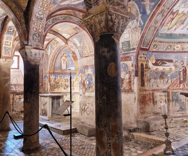 What to see near Rome.Interior of a medieval crypt with frescoed arches, stone columns, and intricate mosaic floors. Description: The image captures the richly decorated interior of an ancient crypt. The vaulted ceiling