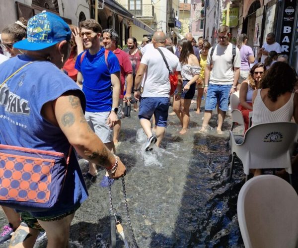 Crowd of people walking barefoot through a shallow stream of water flowing along a narrow Italian street during a summer event, with a man leading a dog on a leash. A Chiena Campagna