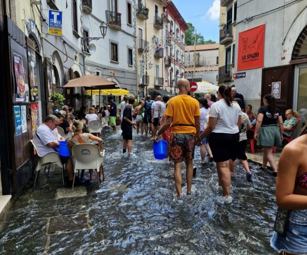 Ludzie brodzący w wodzie na zalanej ulicy podczas święta A Chiena Campagna w Kampanii; niektórzy siedzą w kawiarniach, inni niosą niebieskie wiadra, a w tle widać stare kamienice i wieżę kościoła