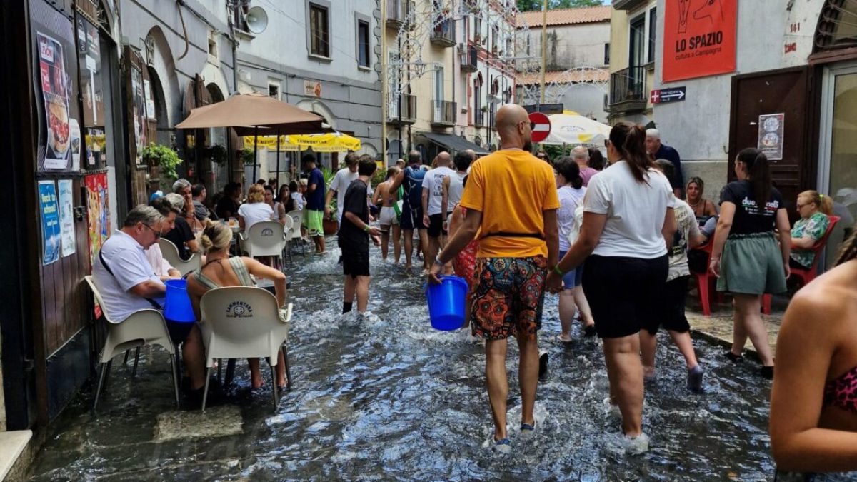 Ludzie brodzący w wodzie na zalanej ulicy podczas święta A Chiena Campagna w Kampanii; niektórzy siedzą w kawiarniach, inni niosą niebieskie wiadra, a w tle widać stare kamienice i wieżę kościoła