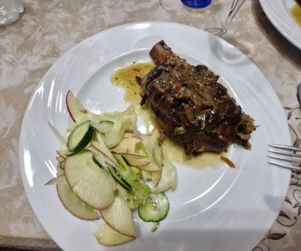 Plate of roasted pork knuckle in sauce served with a fresh side salad of sliced apples, cucumbers, and lettuce on a white tablecloth at Home Restaurant da Elisa in Palombaro, Italy.