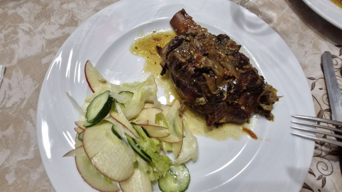 Plate of roasted pork knuckle in sauce served with a fresh side salad of sliced apples, cucumbers, and lettuce on a white tablecloth at Home Restaurant da Elisa in Palombaro, Italy.