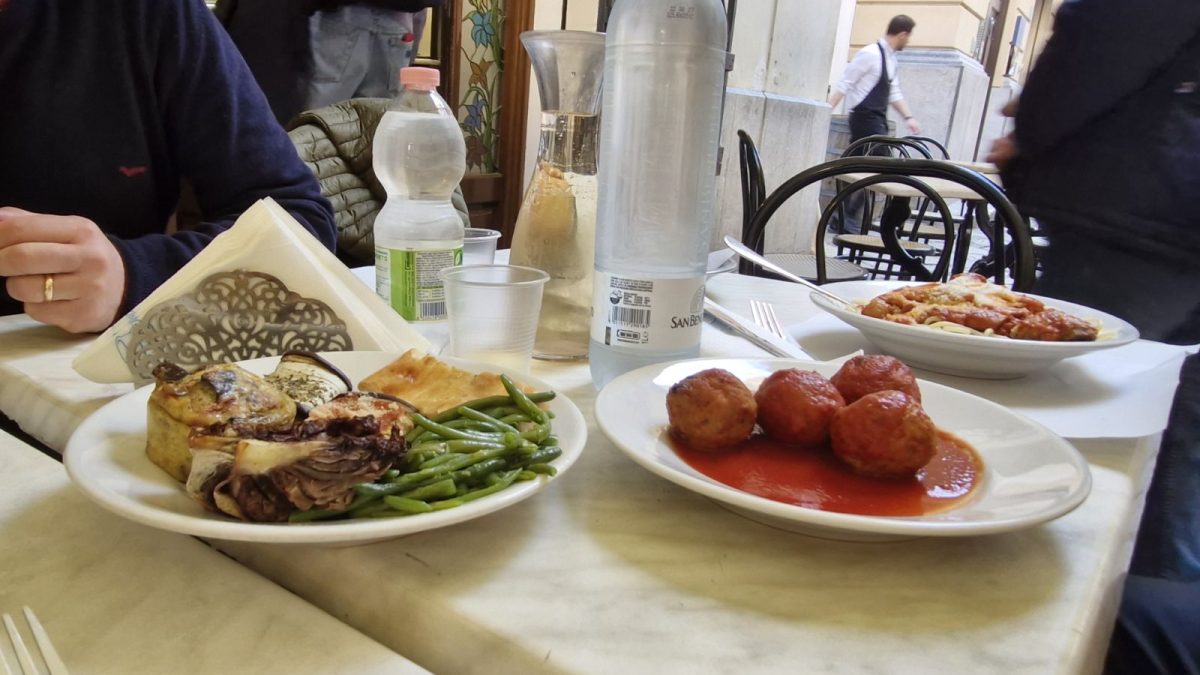 Where locals eat in Palermo. An outdoor restaurant table in Palermo featuring plates of traditional Sicilian food, including large meatballs in tomato sauce, a pasta dish, and a plate with green beans, grilled vegetables, and a slice of focaccia. Two bottles of water and a glass carafe sit on the marble table, with the entrance to Focacceria del Massimo visible in the background.