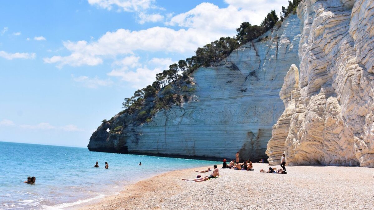 on a pebble Vignanotica Beach under a white cliff covered with trees, several people are sitting on a blanket, three more people are swimming in the blue sea, only their heads are visible