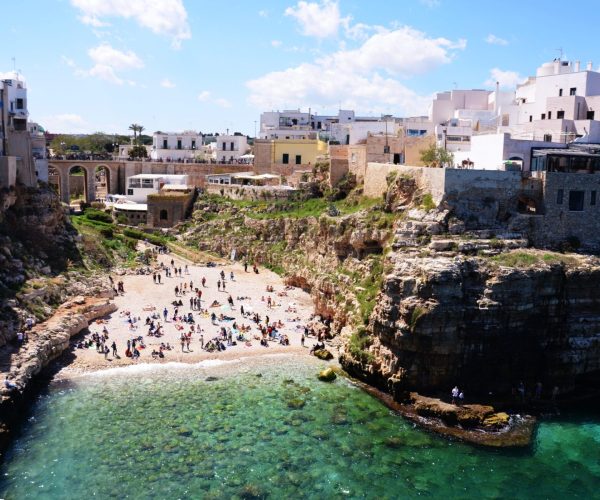 Crowded beach with turquoise water surrounded by cliffs and whitewashed houses in Polignano a Mare, Puglia, Italy.