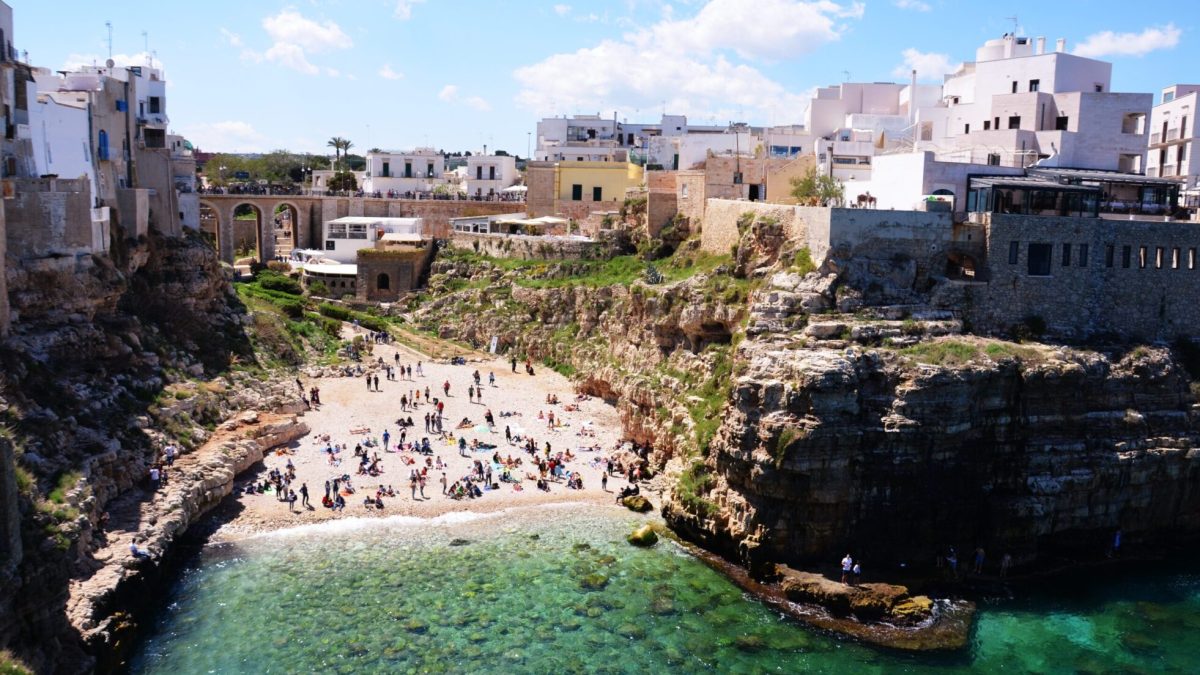 Crowded beach with turquoise water surrounded by cliffs and whitewashed houses in Polignano a Mare, Puglia, Italy.