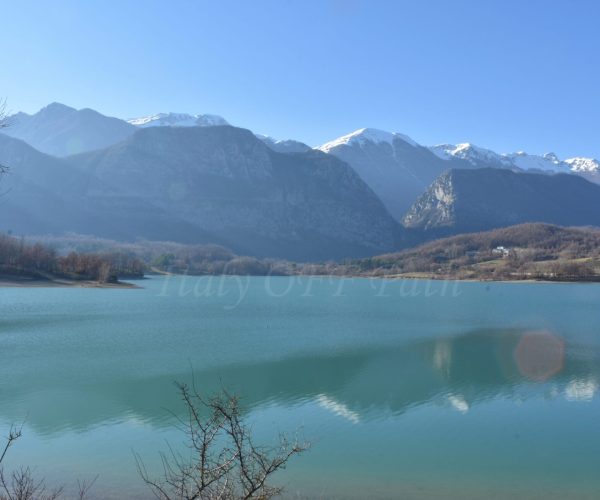 Molise in winter, Clear turquoise lake surrounded by bare winter trees, with snow-capped mountains in the background under a bright blue sky.