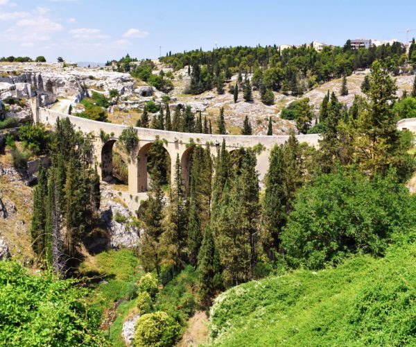 Ancient stone aqueduct surrounded by cypress trees in Gravina di Puglia, Italy, with rocky cliffs and historic buildings in the background in Gravina in Puglia