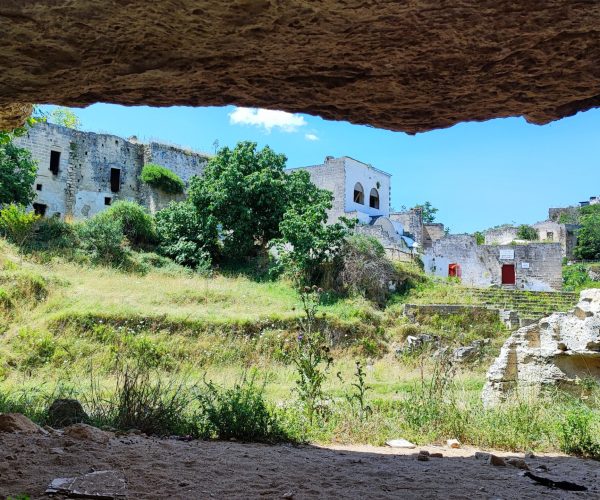 View from inside a cave looking out onto a sunlit grassy valley with old stone buildings and ruins in the background, surrounded by trees and rocky cliffs under a bright blue sky in Gravina di Ginosa