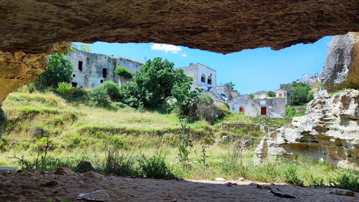 View from inside a cave looking out onto a sunlit grassy valley with old stone buildings and ruins in the background, surrounded by trees and rocky cliffs under a bright blue sky in Gravina di Ginosa