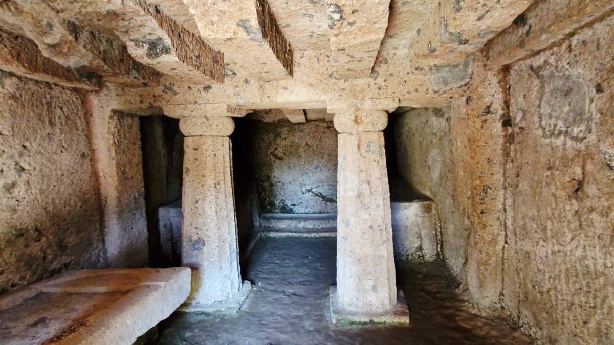 Hikes near Rome. The dark interior of the ancient Etruscan Tomba Margaretha, showing two hand-carved stone columns supporting a rock ceiling with carved beams and funeral stone platforms.