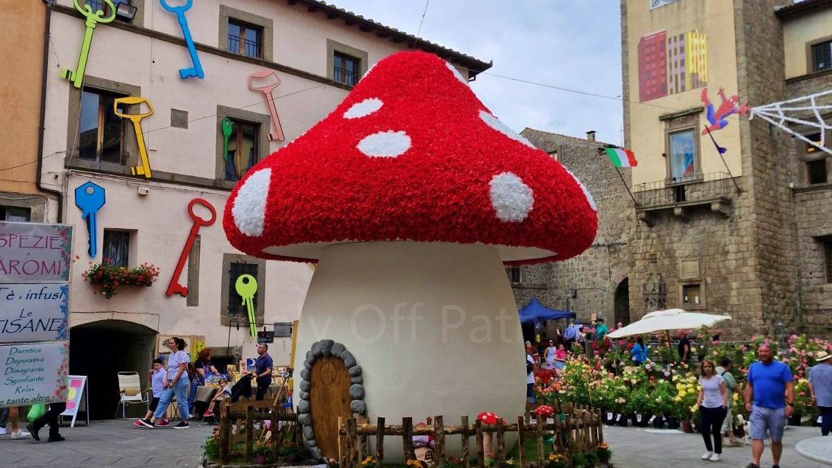 Local festivals in Italy. A large, mushroom-shaped house decoration resembling a red and white fly agaric sits in the center of a stone-paved main square during the Peperino in Fiore festival. The surrounding historic stone buildings are decorated with colorful oversized keys on the walls, and a tall stone clock tower stands in the background under a cloudy sky.