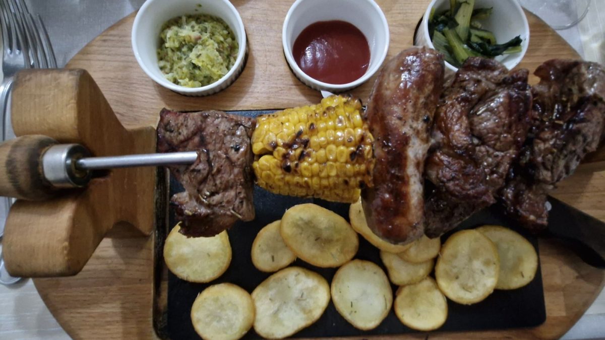 Monti Azzurri Montemonaco. A rustic meat platter served on a wooden board at Ristorante Borgo Antico, featuring a metal skewer with grilled beef, sausage, and corn on the cob over a bed of roasted potato slices, accompanied by three small white bowls containing sauces and cooked chicory