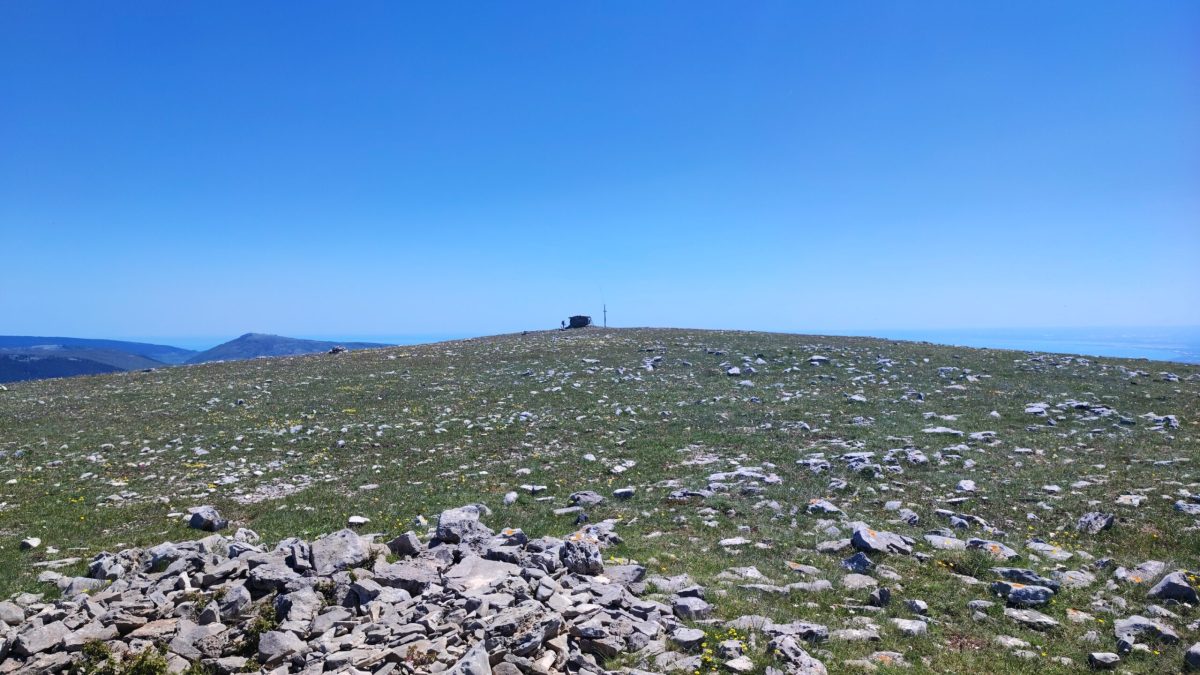 Monte Calvo Gargano, the highest peak in Gargano, featuring the stone refuge and cross against a clear blue sky. The foreground shows the karst landscape with scattered limestone rocks and wild herbs typical of the Puglia highlands.
