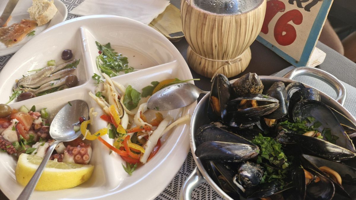 A multi-compartment white ceramic dish containing marinated anchovies, an octopus and tomato salad with lemon, and a squid salad with peppers, served next to a small metal pot filled with steamed mussels at La Sbirra restaurant. La Sbirra Termoli review.
