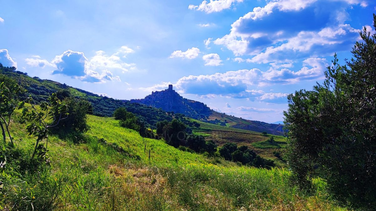 Distant view of the ghost town of Craco in Basilicata, Italy, perched on a hilltop surrounded by green rolling hills and a bright blue sky with scattered clouds