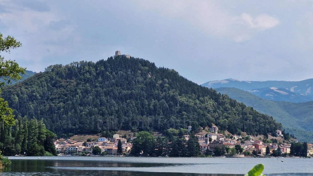 View across Lake Piediluco toward the village at the base of the forested hill with the Rocca Albornoziana ruins at the peak, Umbria, Italy.